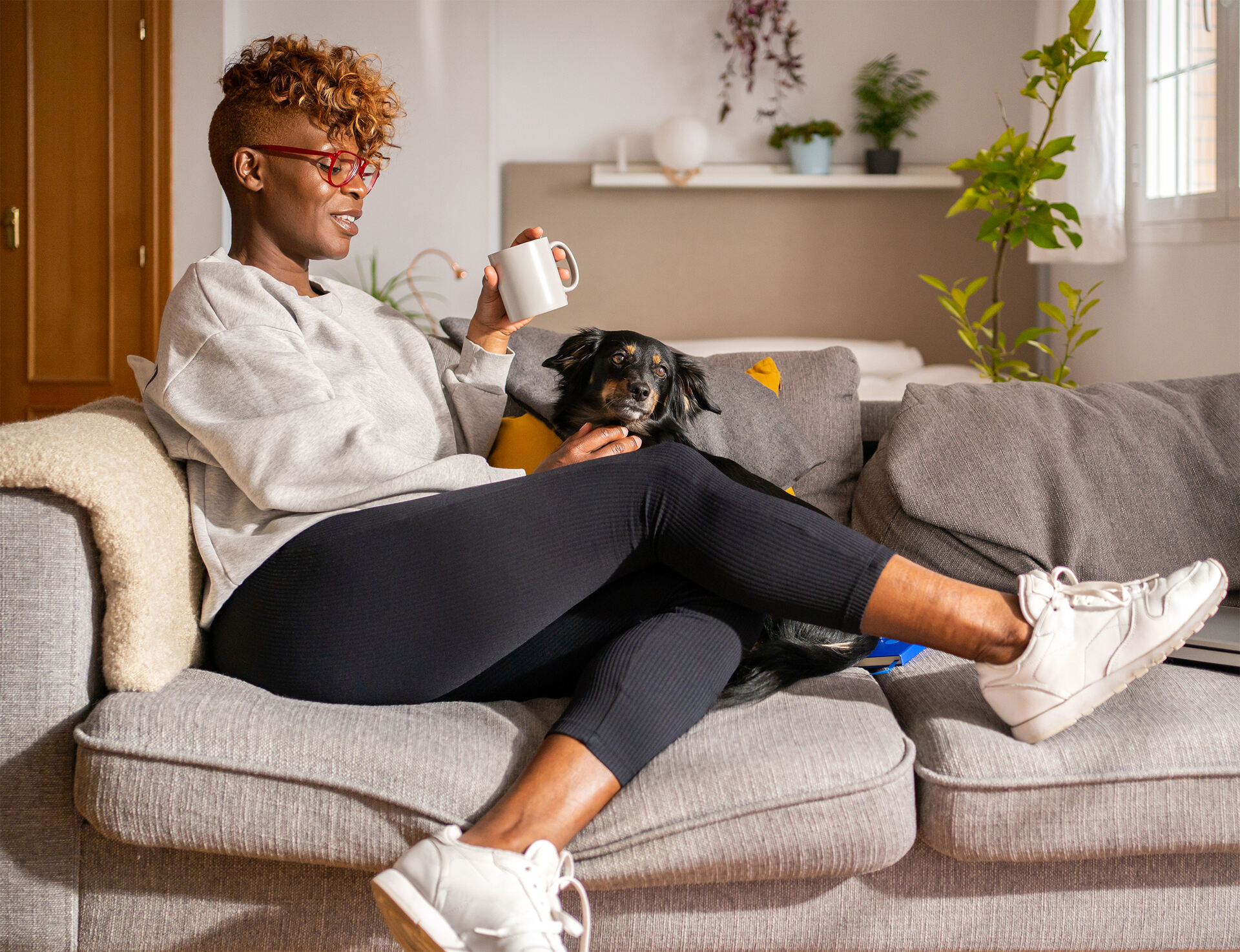 A Black woman in athletic wear drinking coffee on the couch with her dog in a bright modern apartment