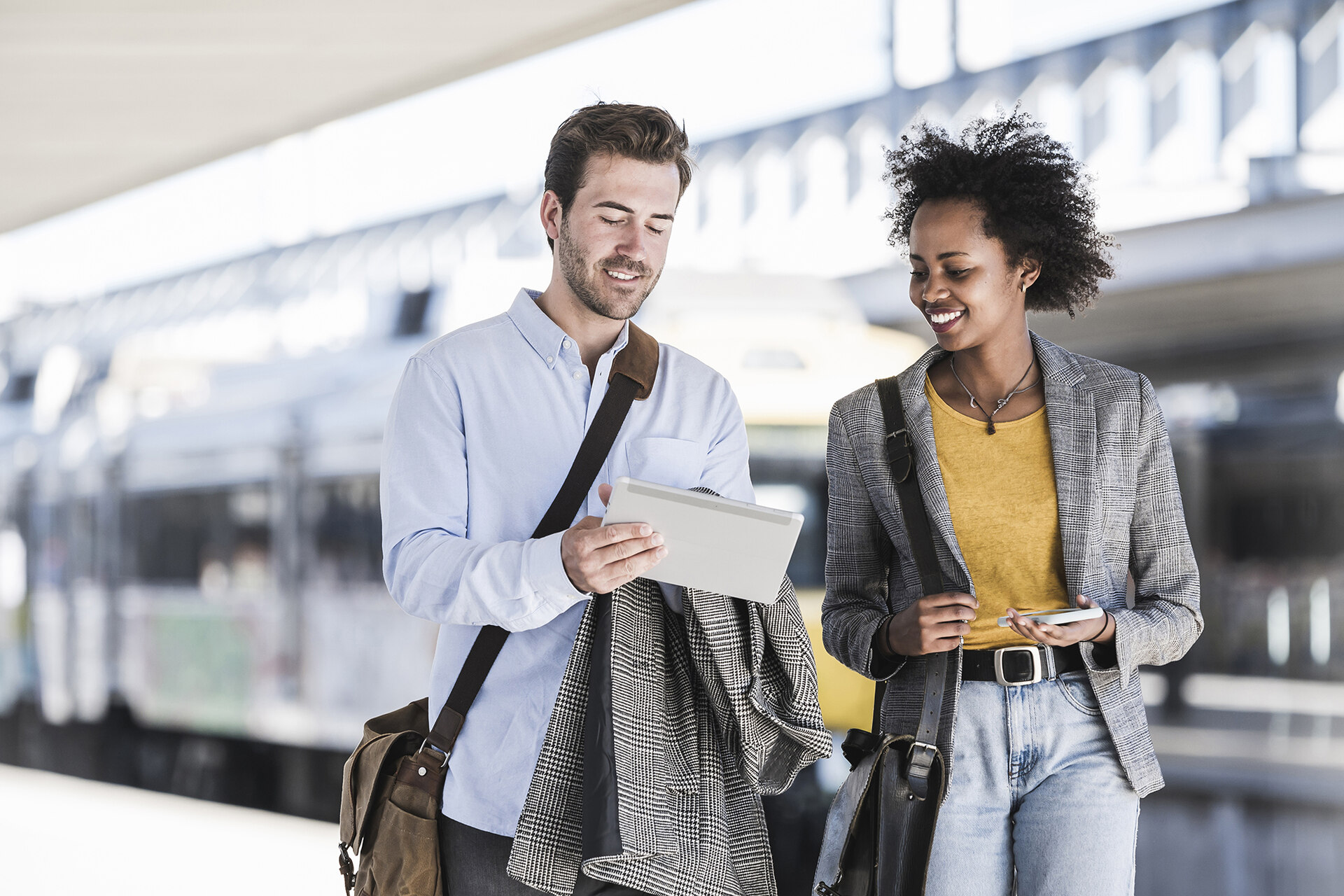 A Black woman and white man in business casual attire looking at a tablet on a train station platform