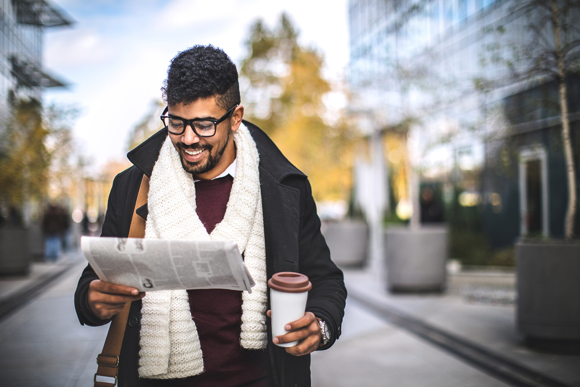 A smiling professional man of color walking down the street reading a newspaper and carrying a to-go coffee