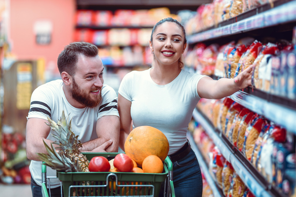 A young couple shopping in a fully-stocked, brightly lit grocery store, pushing a cart holding fresh fruit