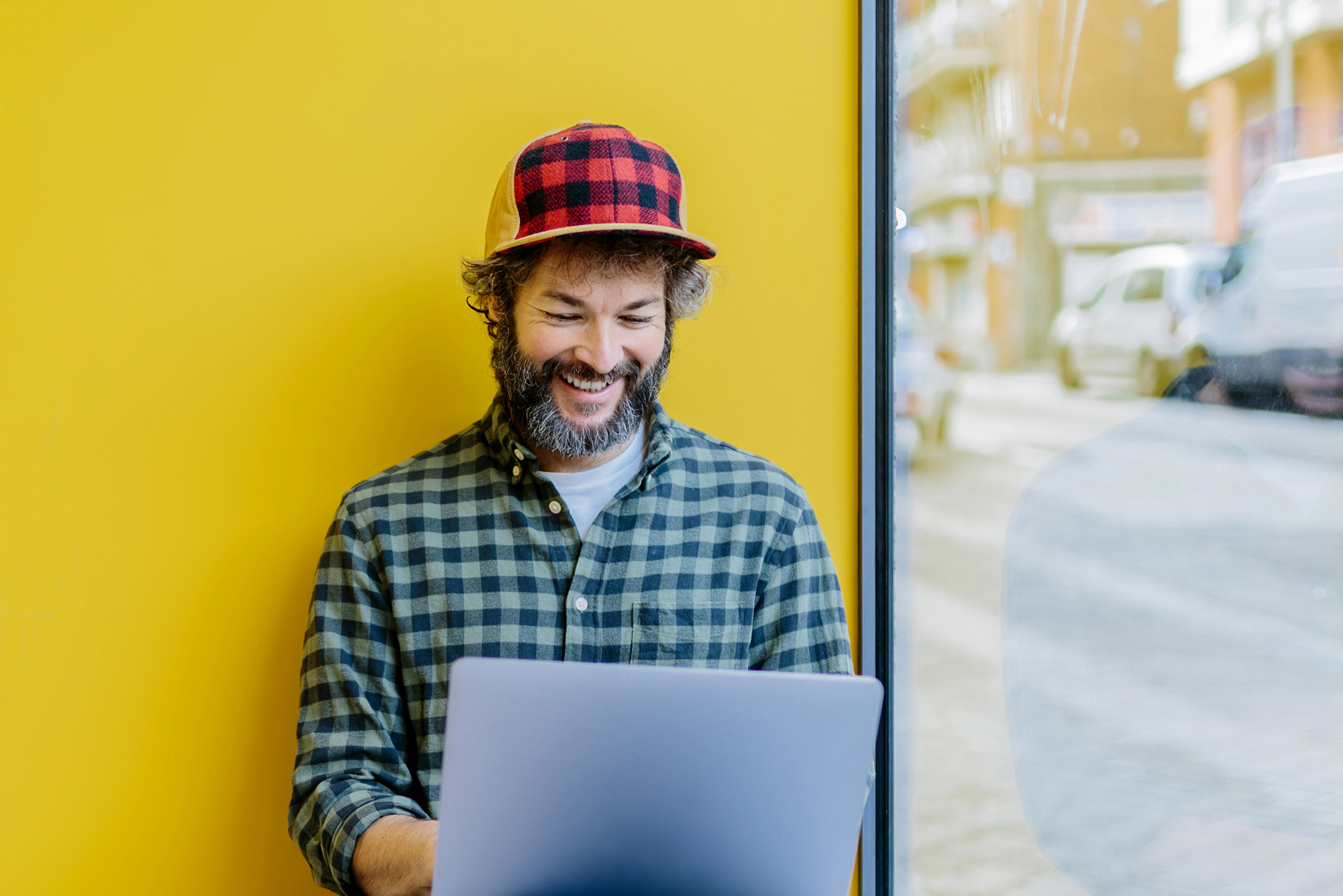 A smiling man working on a laptop against a bright yellow wall next to a large window