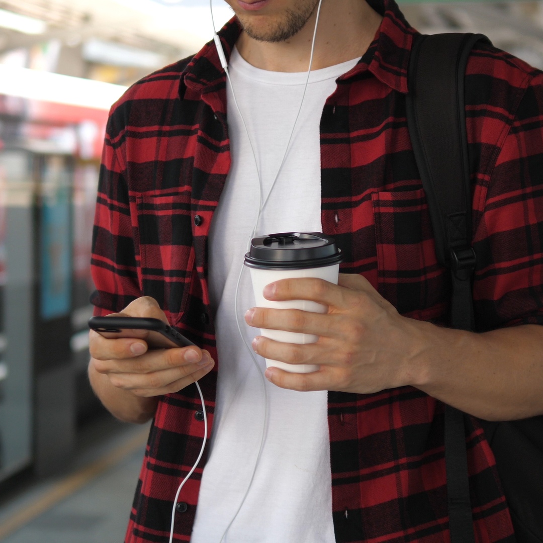 A young man holding a cup of coffee and using his phone while waiting at the train station